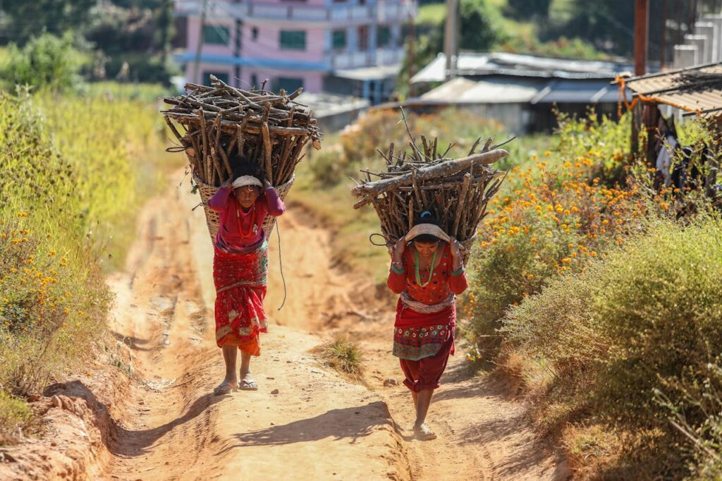 Two women in traditional attire carrying firewood in a rural Nepali landscape.